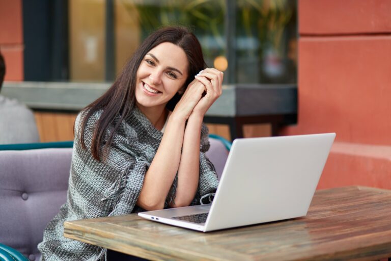 Happy female freelancer satisfied with distance job, works in outdoor restaurant, has coverlet on shoulders, sits in front of opened laptop, looks happily, models alone. People, leisure and technology