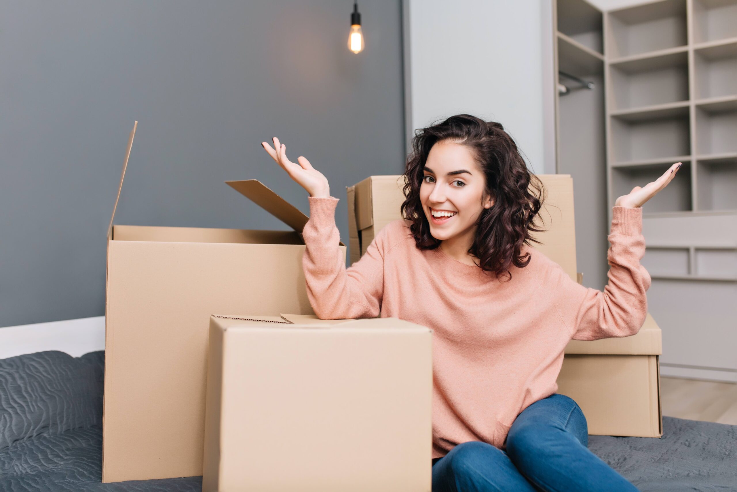 Excited young woman on bed surround boxes, carton smiling to camera in modern apartment. Moving to new flat, expressing true positive emotions at new home with modern interior